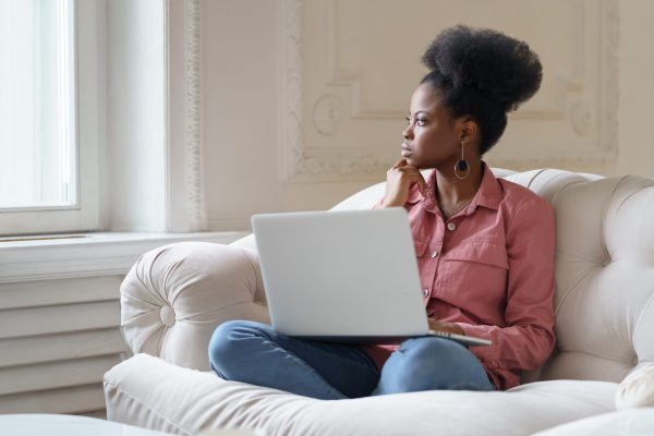 Thoughtful serious African American millennial woman on couch at home, on laptop.