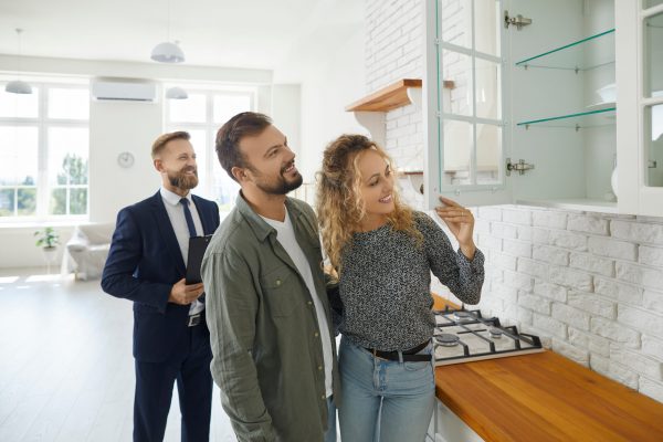 Boyfriend and girlfriend buying house. First time buyers or future tenants meeting real estate agent and looking around new home. Husband and wife looking at good quality modern wooden kitchen cabinet